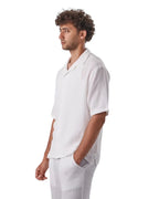 A man wearing a white My Store linen short set with a short-sleeved button-down shirt and matching shorts.